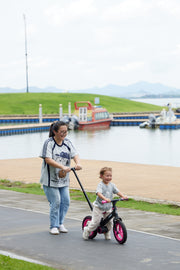 Parent guiding a smiling toddler riding a pink and black JMMD S900 kids trike by the lakeside